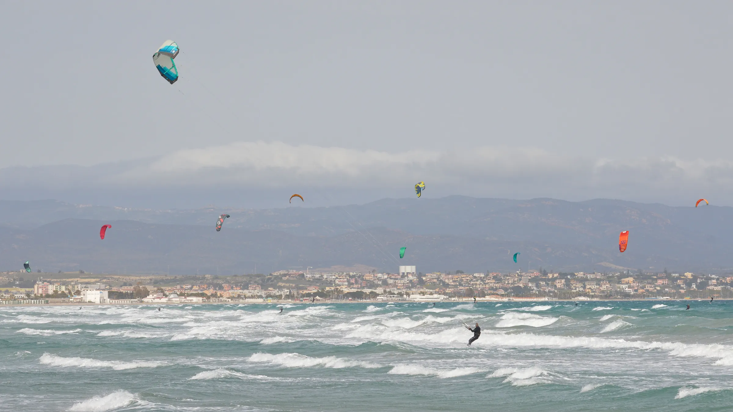 Kitesurfers met kleurrijke kites boven de golven in de Golf van Cagliari, met de stad en de bergen op de achtergrond