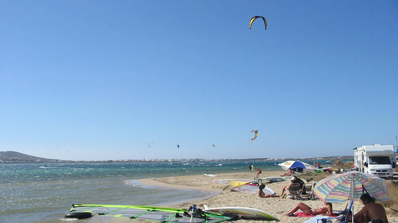 Kites boven de zeestraat bij Pounda op Paros, met windsurfspullen op het strand